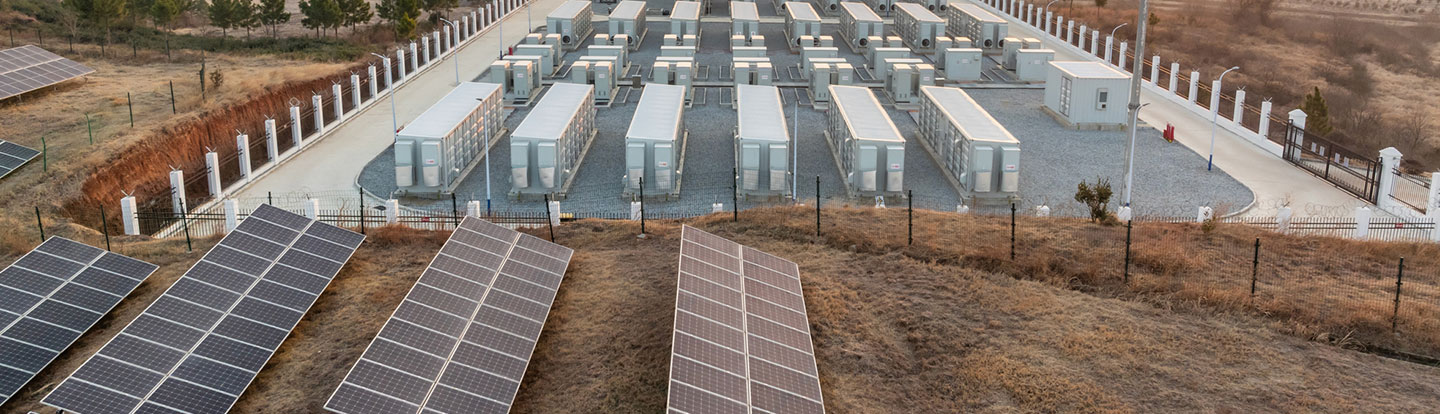 Vista aerea di un impianto di pannelli solari con batterie di accumulo energetico disposte in file ordinate, in un’area recintata su terreno brullo.