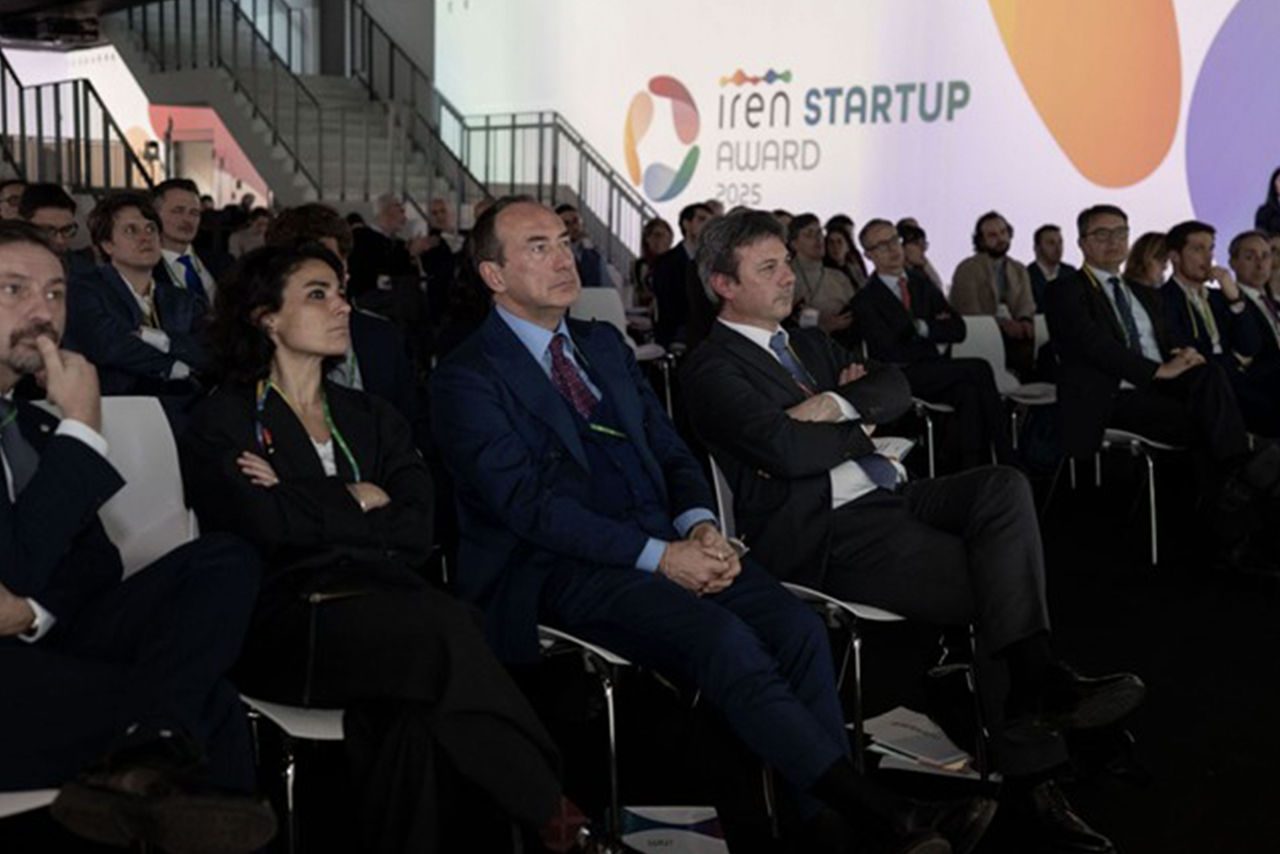 Pubblico attento durante la cerimonia degli Iren Startup Awards 2025, con il logo dell'evento proiettato sulla parete. In primo piano, dirigenti e professionisti in abiti formali ascoltano con interesse.