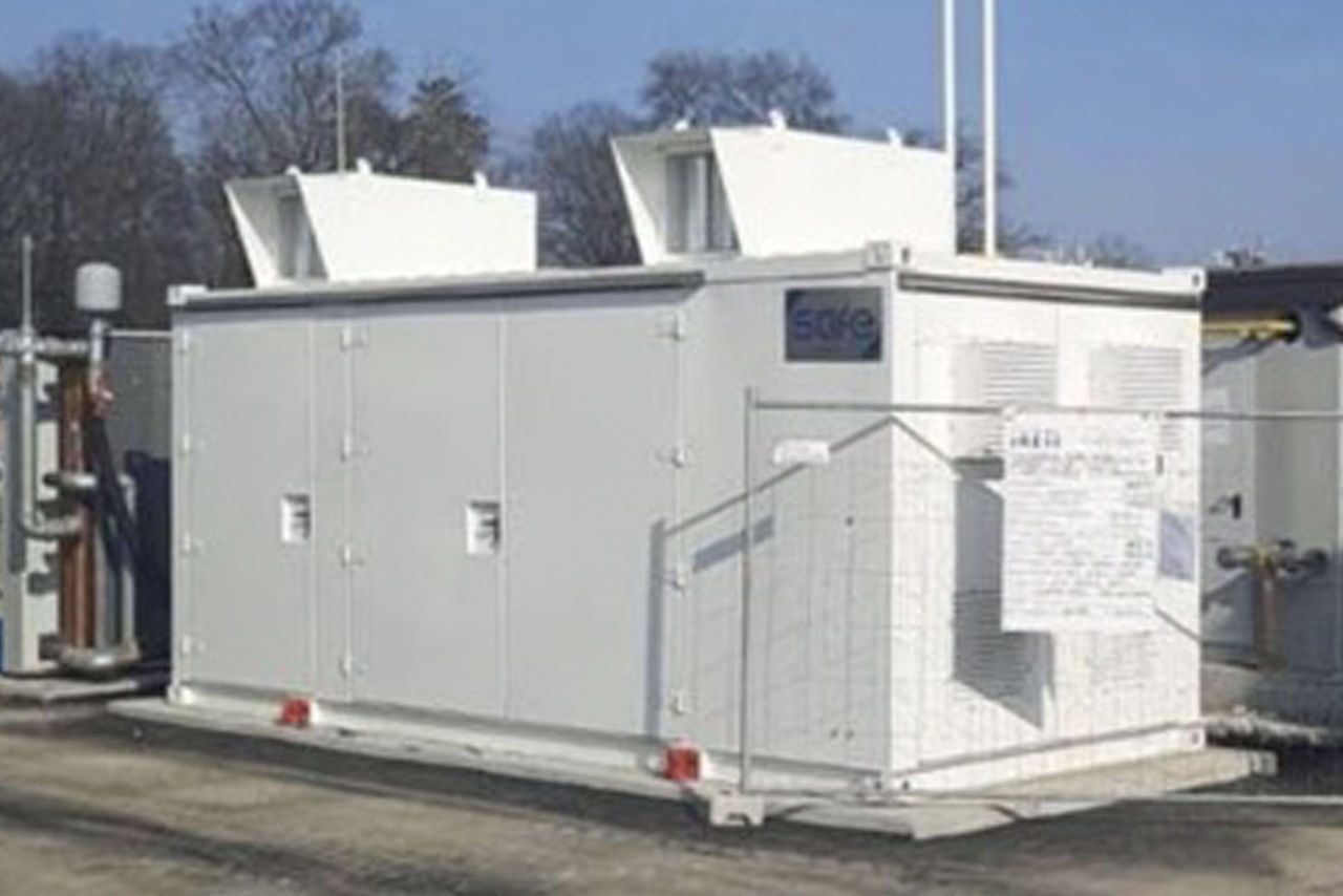 Struttura bianca prefabbricata situata in un'area industriale, parte di un impianto per la gestione del gas o dell'energia, circondata da recinzioni e con elementi tecnici visibili sul tetto.