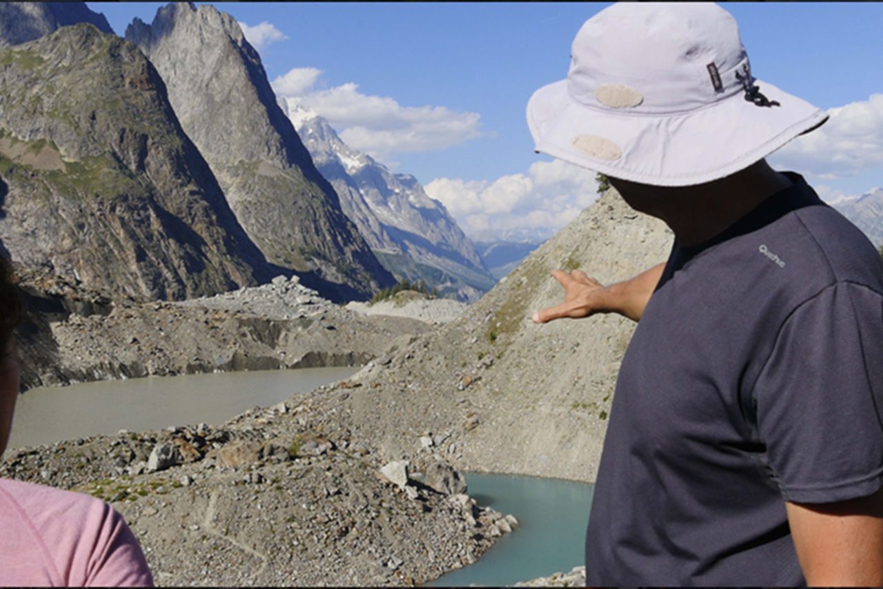uomo di spalle con cappellino che indica il fiume e le montagne all'orizzonte nell'atto di spiegare