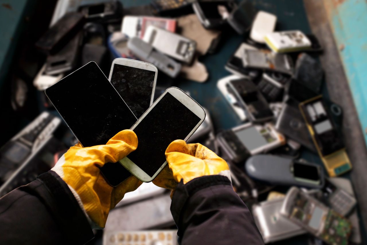 Persona con guanti gialli che tiene tre smartphone usati davanti a un cumulo di dispositivi elettronici dismessi