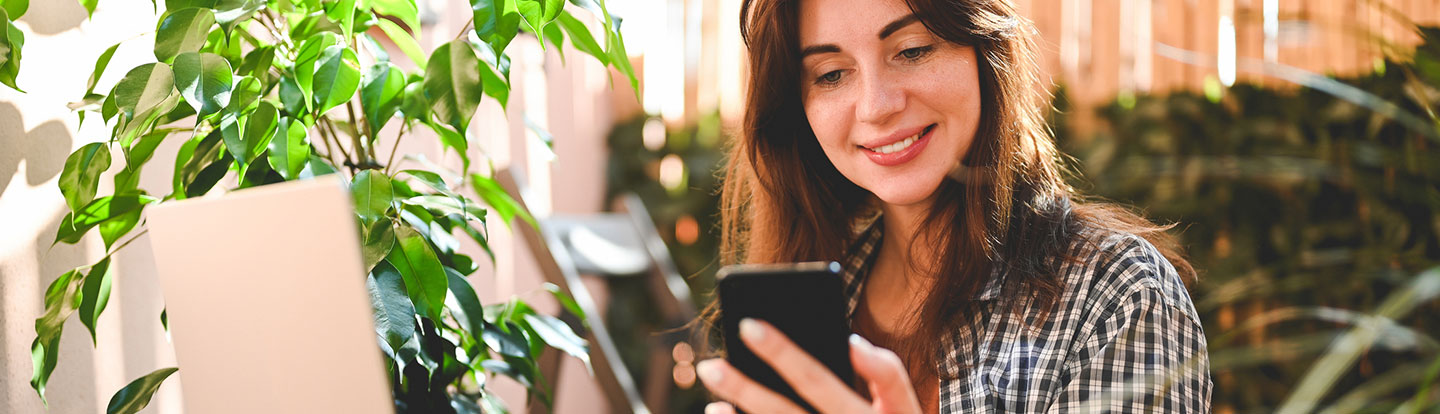 Donna sorridente seduta con lo smartphone in mano accanto a piante verdi in un ambiente domestico luminoso