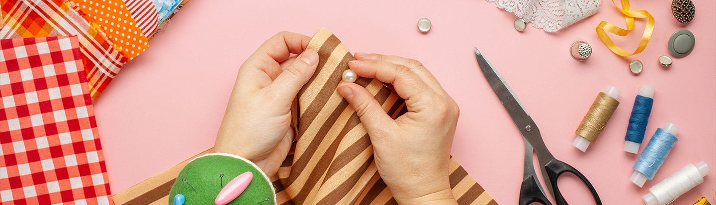 Mani che cuciono una perla su un tessuto a righe, circondate da forbici, bottoni, pizzi e materiali da cucito su sfondo rosa, a rappresentare il riuso creativo e la sartoria sostenibile.