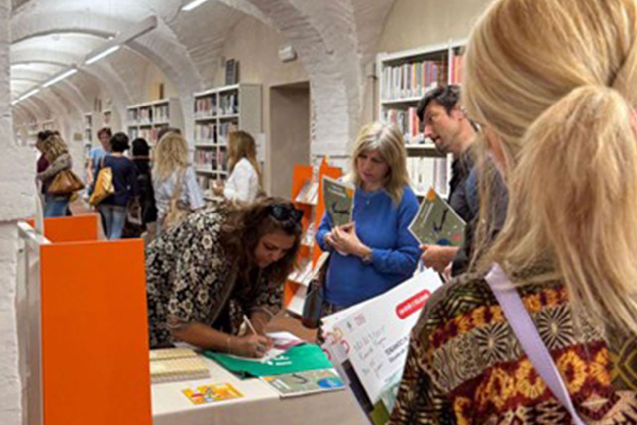 Persone consultano materiali informativi presso uno stand Eduiren allestito all’interno di una biblioteca con archi in mattoni bianchi.