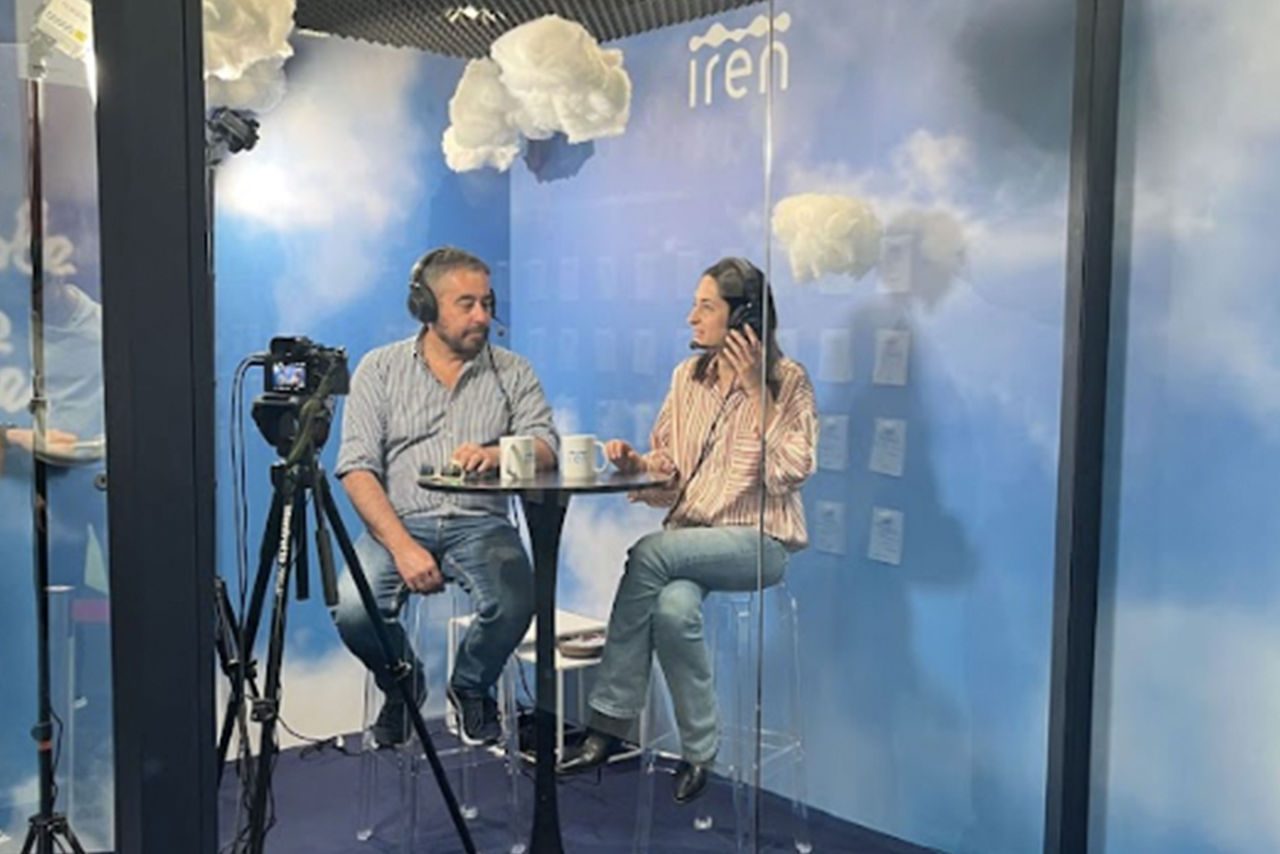 Due persone all'interno di uno studio in vetro con sfondo azzurro e nuvole finte, sedute a un tavolo con microfoni e cuffie per registrare un podcast a tema "Le parole che vorrei" di Iren.