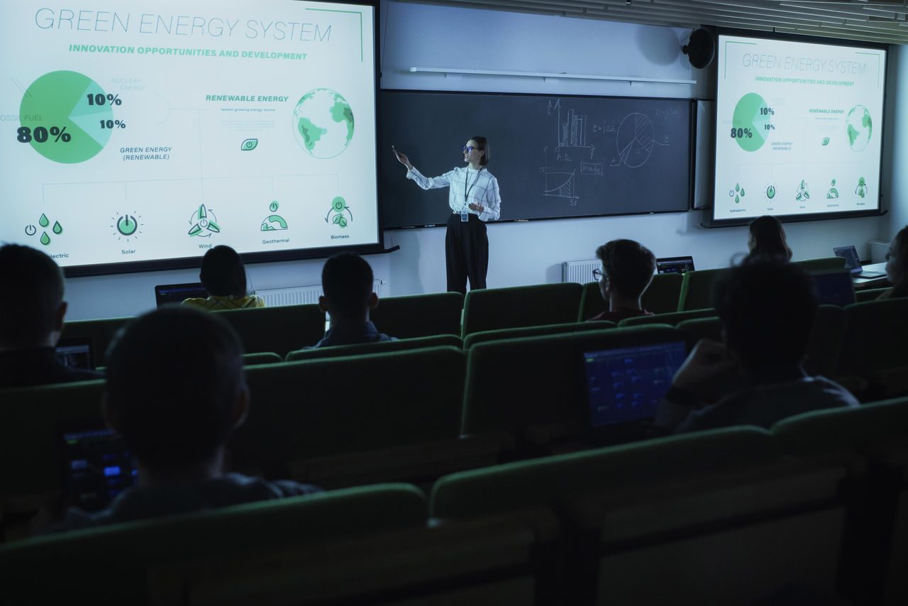 in primo piano di spalle studenti seduti in aula ad anfiteatro e in secondo piano in cattedra una docente che tiene la lezione indicando il proiettore con dati sul green energy system